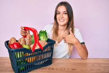 Obraz na płótnie Canvas Beautiful caucasian woman holding supermarket shopping basket smiling happy pointing with hand and finger