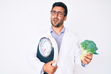 Young hispanic man as nutritionist doctor holding weighing machine and broccoli clueless and confused expression. doubt concept.
