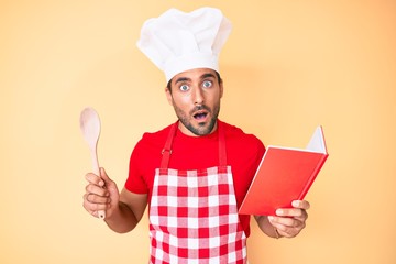 Young hispanic man wearing professional baker apron reading cooking recipe book in shock face,...