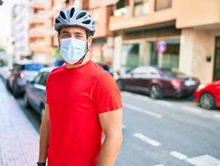 Young delivery man wearing bike helmet and coronavirus protection medical mask standing at town street.