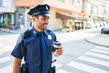 Young hispanic policeman wearing police uniform smiling happy. Drinking cup of take away coffee standing at town street.