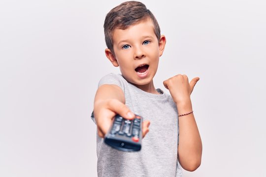 Cute Blond Kid Holding Television Remote Control Pointing Thumb Up To The Side Smiling Happy With Open Mouth