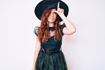 Young beautiful woman wearing witch halloween costume making fun of people with fingers on forehead doing loser gesture mocking and insulting.