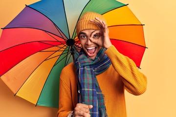 Young african amercian man holding colorful umbrella smiling happy doing ok sign with hand on eye looking through fingers
