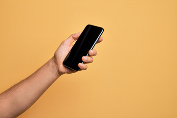 Hand of caucasian young man holding smartphone showing screen over isolated yellow background