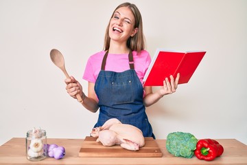 Young blonde girl wearing professional baker apron reading cooking recipe book winking looking at...