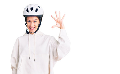 Beautiful brunette young woman wearing bike helmet and sporty clothes smiling and confident gesturing with hand doing small size sign with fingers looking and the camera. measure concept.