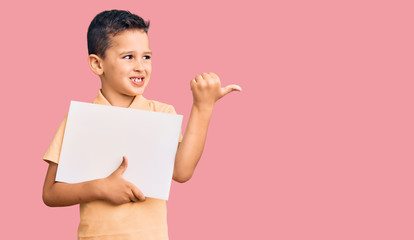 Little cute boy kid holding cardboard banner with blank space pointing thumb up to the side smiling happy with open mouth