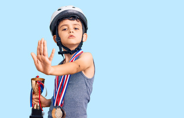 Little cute boy kid wearing bike helmet and winner medals holding winner trophy with open hand doing stop sign with serious and confident expression, defense gesture