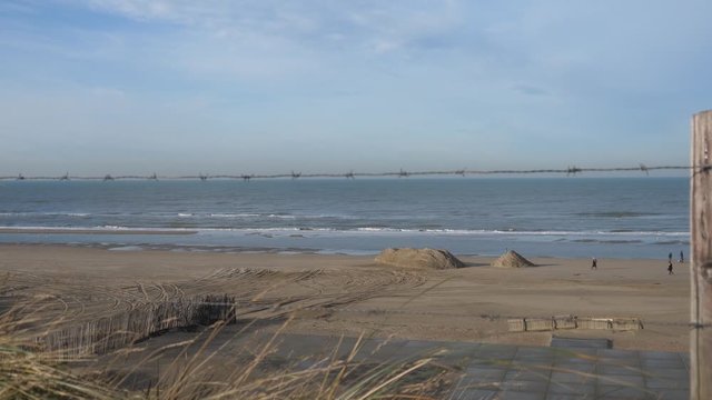 In the out of focus foreground a barbed wire fence and grass. View of the cleared empty winter beach in Zandvoort Netherlands, from an elevated position. Few strollers, surf and water of the North Sea