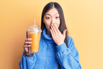 Young beautiful chinese woman drinking glass of orange juice covering mouth with hand, shocked and...
