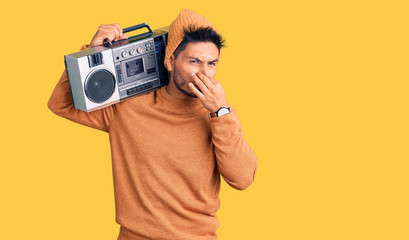 Handsome latin american young man holding boombox, listening to music smelling something stinky and disgusting, intolerable smell, holding breath with fingers on nose. bad smell