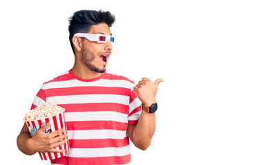 Handsome latin american young man wearing 3d glasses and eating popcorn at the movies pointing thumb up to the side smiling happy with open mouth