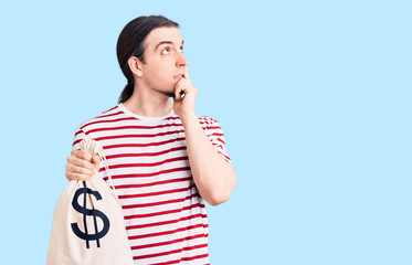 Young adult man with long hair holding money bag with dollar symbol serious face thinking about...