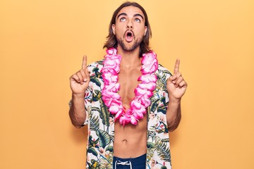 Young handsome man wearing swimwear and hawaiian lei amazed and surprised looking up and pointing...
