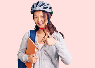 Young beautiful chinese girl wearing student backpack and bike helmet holding book smiling happy pointing with hand and finger