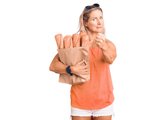 Middle age fit blonde woman holding paper bag with bread annoyed and frustrated shouting with anger, yelling crazy with anger and hand raised