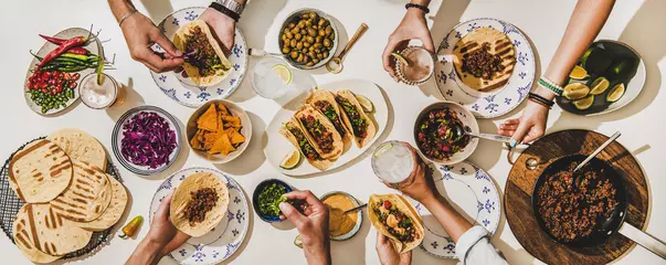 Photo sur Plexiglas Manger Amis ayant un dîner de tacos mexicains. Mise à plat de tacos au bœuf, salsa à la tomate, tortillas, bière, collations et mains des peuples sur une table blanche, vue de dessus. Cuisine mexicaine, rassemblement, fête, concept de nourriture réconfortante