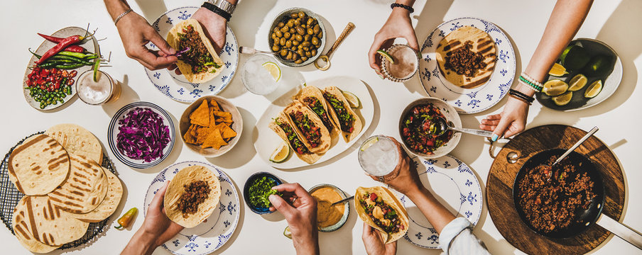 Friends having Mexican Taco dinner. Flat-lay of beef tacos, tomato salsa, tortillas, beer, snacks and peoples hands over white table, top view. Mexican cuisine, gathering, feast, comfort food concept