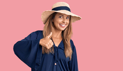 Middle age hispanic woman wearing summer hat doing happy thumbs up gesture with hand. approving expression looking at the camera showing success.