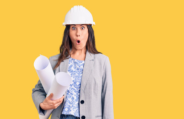 Young beautiful brunette woman wearing architect hardhat holding blueprint scared and amazed with open mouth for surprise, disbelief face
