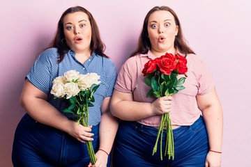 Young plus size twins holding flowers scared and amazed with open mouth for surprise, disbelief face
