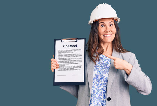 Young beautiful brunette woman wearing architect hardhat holding contract smiling happy pointing with hand and finger