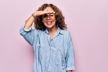 Middle age beautiful woman wearing casual denim shirt standing over pink background very happy and smiling looking far away with hand over head. Searching concept.