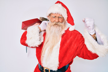 Old senior man with grey hair and long beard wearing santa claus costume holding shopping bags annoyed and frustrated shouting with anger, yelling crazy with anger and hand raised