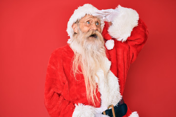 Old senior man with grey hair and long beard wearing traditional santa claus costume very happy and smiling looking far away with hand over head. searching concept.