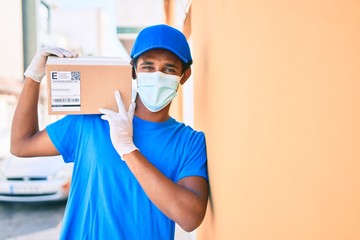 Fototapeta na wymiar African delivery man wearing courier uniform outdoors wearing coronavirus safety mask holding cardboard parcel