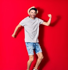 Fototapeta premium Young handsome hispanic man wearing casual clothes and summer hat smiling happy. Jumping with smile on face celebrating with fists up over isolated red background.