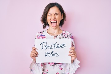 Young beautiful girl holding positive vibes banner sticking tongue out happy with funny expression.