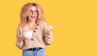 Young blonde woman with curly hair wearing glasses and drinking a cup of coffee pointing thumb up to the side smiling happy with open mouth