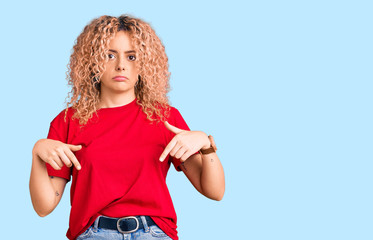 Young blonde woman with curly hair wearing casual red tshirt pointing down looking sad and upset, indicating direction with fingers, unhappy and depressed.