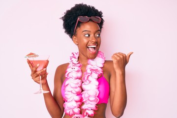 Young african american woman wearing bikini and hawaiian lei holding cocktail pointing thumb up to...