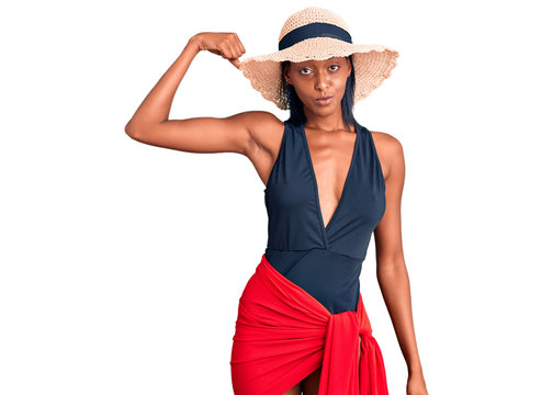 Young african american woman wearing swimsuit and summer hat strong person showing arm muscle, confident and proud of power