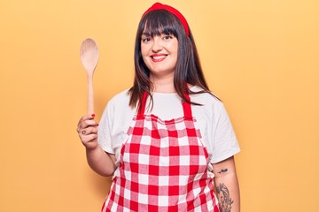 Young plus size woman wearing apron holding wooden spoon looking positive and happy standing and smiling with a confident smile showing teeth