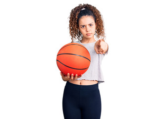 Beautiful kid girl with curly hair holding basketball ball pointing with finger to the camera and to you, confident gesture looking serious