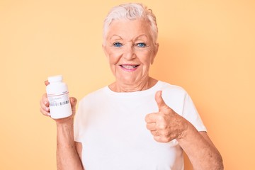 Senior beautiful woman with blue eyes and grey hair holding pills smiling happy and positive, thumb up doing excellent and approval sign