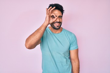 Handsome hispanic man with beard wearing casual clothes doing ok gesture with hand smiling, eye looking through fingers with happy face.