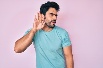 Handsome hispanic man with beard wearing casual clothes smiling with hand over ear listening an hearing to rumor or gossip. deafness concept.
