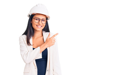 Young beautiful latin girl wearing architect hardhat and glasses cheerful with a smile of face pointing with hand and finger up to the side with happy and natural expression on face