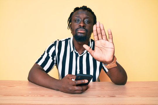 Young african american man with braids using smartphone sitting on the table with open hand doing stop sign with serious and confident expression, defense gesture