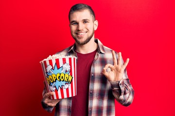 Young handsome man eating popcorn doing ok sign with fingers, smiling friendly gesturing excellent symbol
