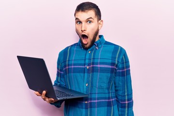 Young handsome man working using laptop scared and amazed with open mouth for surprise, disbelief face