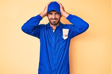 Handsome young man with curly hair and bear wearing builder jumpsuit uniform suffering from headache desperate and stressed because pain and migraine. hands on head.