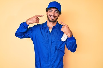 Handsome young man with curly hair and bear wearing builder jumpsuit uniform smiling cheerful showing and pointing with fingers teeth and mouth. dental health concept.