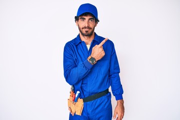 Handsome young man with curly hair and bear weaing handyman uniform pointing with hand finger to the side showing advertisement, serious and calm face