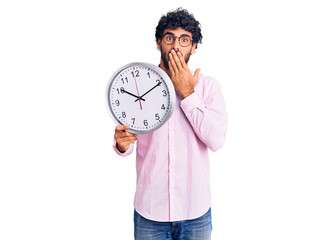 Handsome young man with curly hair and bear holding big clock covering mouth with hand, shocked and...